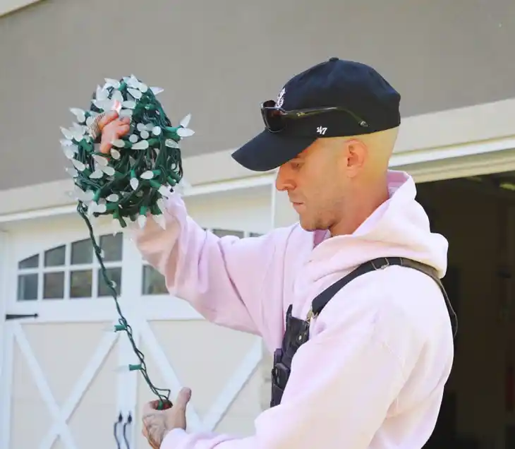 unwrapping-christmas-ligths Man in pink hoodie and black cap untangling a bundle of Christmas lights in preparation for outdoor holiday decorating.